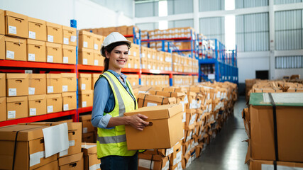 Professional female warehouse worker in safety vest and hard hat holding a cardboard box in a large storage facility. Logistics, inventory management, and industrial supply chain concept.