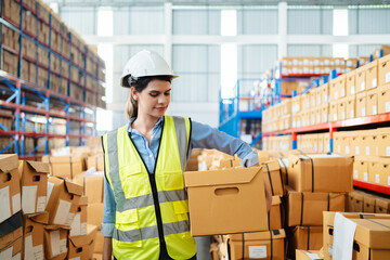 Professional female warehouse worker in safety vest and hard hat holding a cardboard box in a large storage facility. Logistics, inventory management, and industrial supply chain concept.