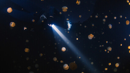 Underwater shot of the freediver swimming with the torch at night in the lake full of jellyfish.