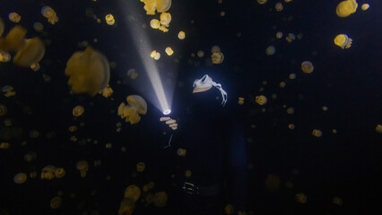 Underwater shot of the freediver swimming with the torch at night in the lake full of jellyfish.