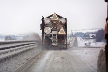 Obraz premium Winter road maintenance truck spreads deicing salt on rural icy highway, warning triangles glow, snow spray trails behind.