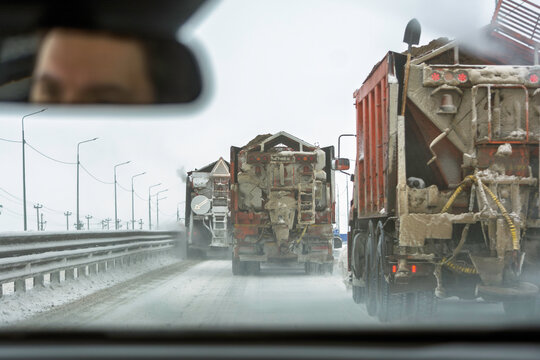 Windshield view shows convoy of salt spreader trucks on snowy highway, rearview mirror shows cautious driver in cold winter.