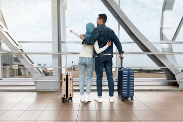 Fototapeta premium Weekend Travel. Loving Islamic Couple Standing Near Window In Airport And Pointing Away, Rear View Of Muslim Spouses With Suitcases Waiting For Flight At Terminal, Enjoying Upcoming Trip, Copy Space