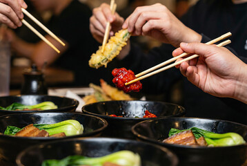 Obraz premium Close-up of friends eating Asian food with chopsticks including tempura and braised pork belly bowls with bok choy in a restaurant