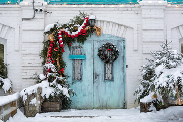 Snowy turquoise wooden door with Christmas wreath and garland on white brick facade, festive winter entrance for holiday decor themes.