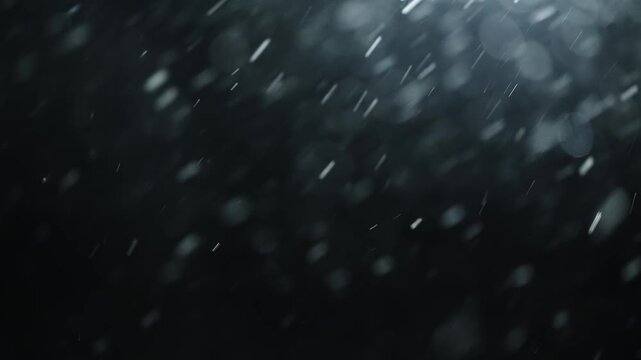 Backlit snow particles drifting through darkness with shimmering bokeh, captured in slow motion as delicate precipitation falls against a black background, evoking winter calm