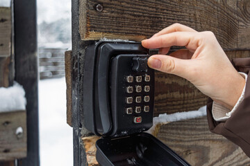 Hand sets code on outdoor keypad lockbox on wooden gate in snowy winter, secure key access for cabin rental self check in.