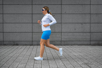 Beautiful middle-aged woman running against background of concrete wall in city on summer day. Side view	