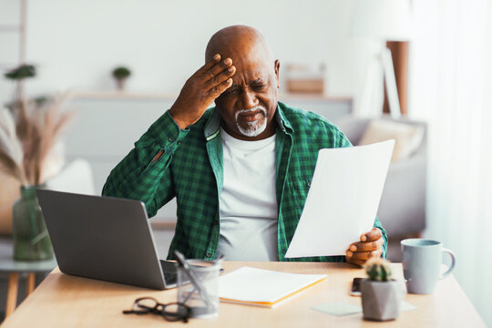 An older man sits at a home office desk, looking worried as he holds paperwork. A laptop is open in front of him, and he appears to be under pressure.