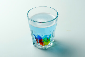 Rainbow child orthodontic retainer rests in blue cleaning solution inside clear glass, calm hygiene concept on white background.
