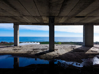 View of the ocean from an unfinished building during daytime with cloudy skies and concrete pillars framing the scene along a coastal area