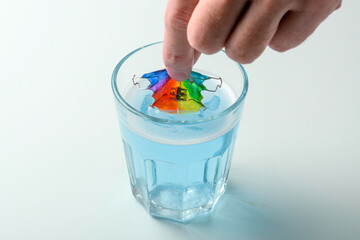 Adult hand places rainbow child orthodontic retainer into glass of blue cleansing solution, careful daily care on light.