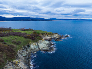 Seaside view from a rocky cliff during a cloudy day near a coastal town in the distance showcasing the waves and landscape