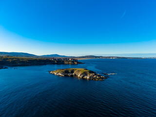 Coastal scene shows calm waters and green land under clear blue sky during sunny day near the shore with distant hills in the background