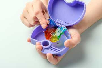 Child places glitter rainbow retainer inside purple travel case, simple routine for hygienic transfer after orthodontic treatment.