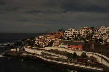 Coastal village with white residential buildings and terracotta roofs perched on a rocky shoreline above the ocean. Dark clouds gather over the sea, creating a dramatic contrast.