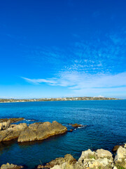 Scenic view of a clear blue ocean and rocky shoreline during daytime with a distant land visible under a bright sky