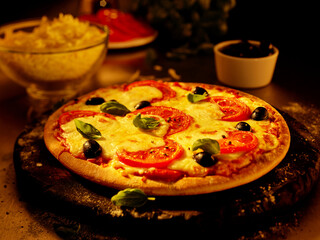  Pizza Margherita mit Oliven und Basilkumbl&auml;ttern auf rustikalem rundem Holzbrett mit gehobeltem K&auml;se in Glasschale