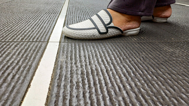 Side view of a person wearing white mules shoes with black accents walking on textured gray tiles. Fashionable footwear, urban lifestyle, and casual street style concept.