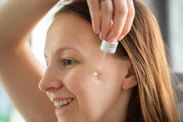 Young woman performing skincare routine at home, applying facial serum and brushing hair. Self-care, beauty and wellness concept.