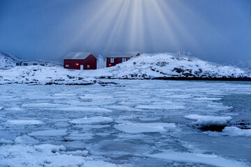 Red wooden houses stand along a frozen coastal shoreline with drifting ice and snow-covered mountains in the background. The quiet winter scene captures the cold, remote atmosphere of a Nordic or Arct © murattellioglu
