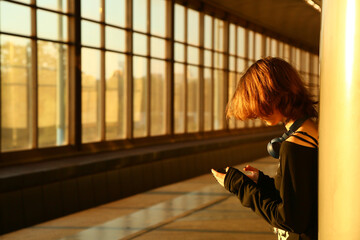 Teen girl waiting in a subway station with windows in sunlight. Young woman standing against a column in style clothes, looking in her mobile phone, natural person pose. Indoors perspective closeup