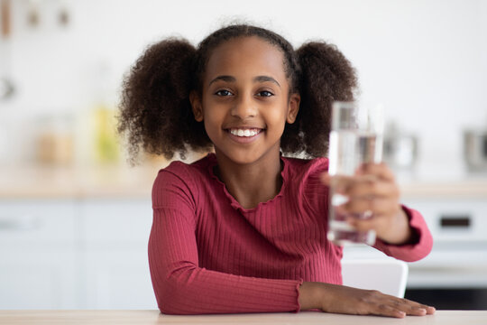 Cheerful african american pretty girl teenager holding glass of fresh water while sitting at table, smiling at camera, kitchen interior, copy space. Hydration, spring water drinking for kids concept