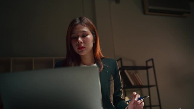 Young Asian business woman working late in a dark office, reviewing documents and using her laptop, finally smiling with satisfaction