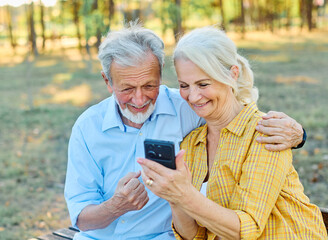 Happy active senior couple having fun using smart phones sitting on a bench outdoors