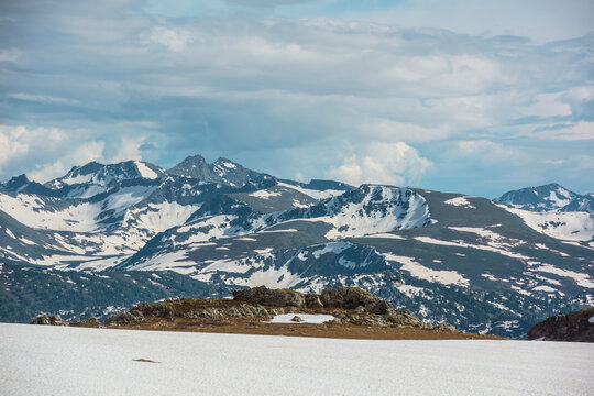 Scenic view from sunlit snow field to gold stone outcrops and sharp golden rocks against pointy peaks and peaked tops under cloudy sky. Wild rugged alpine landscape with big rocky snowy mountain range - Powered by Adobe