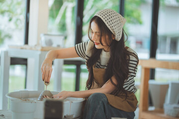 Young Asian woman artisan using a wet sponge to shape and smooth clay on a spinning pottery wheel in a studio