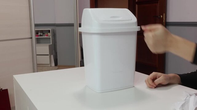 Adult woman hands throwing crumpled paper into white plastic trash bin indoors, symbolizing waste disposal, rejection, cleanup and environmental concept.