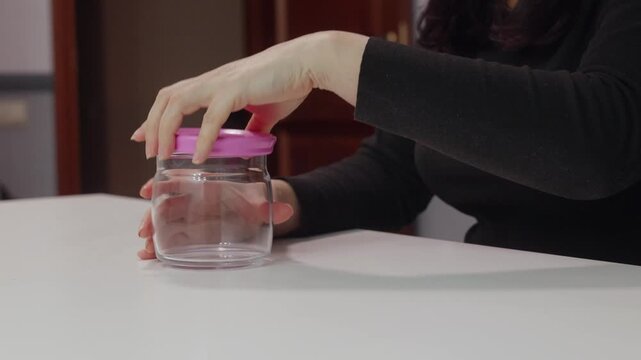 Woman hands closing plastic lid on empty glass jar on white table indoors, illustrating food storage, kitchen organization and reusable container concept.