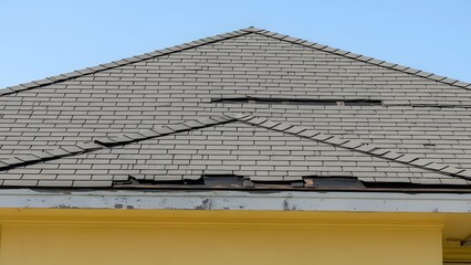 Close up of damaged gray gable roof tiles with yellow exterior wall under clear sky