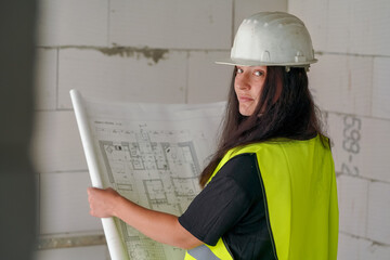 Young female construction worker in green high visibility vest and white hard hat, holding paper drawings with building documentation blueprint, looking over her shoulder, bare walls background