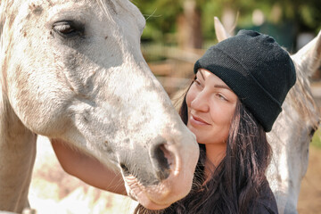 Young woman in beanie hat standing near her white Arabian horse on ranch farm, smiling