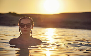 Young woman with sunglasses, standing in evening sea, closeup photo, afternoon sunset background