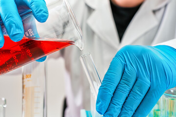 Hands in protective gloves pouring liquid from beaker to a smaller test tube, closeup detail. Chemical lab experiment or analysis