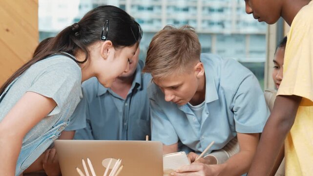 Happy high school boy receive advise and taking a note while caucasian teacher listening and using laptop analyze data. Group of diverse student help teenager solve problem at classroom. Edification