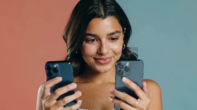 Cheerful young woman comparing two smartphones. Tech enthusiast reviewing mobile devices against split color background