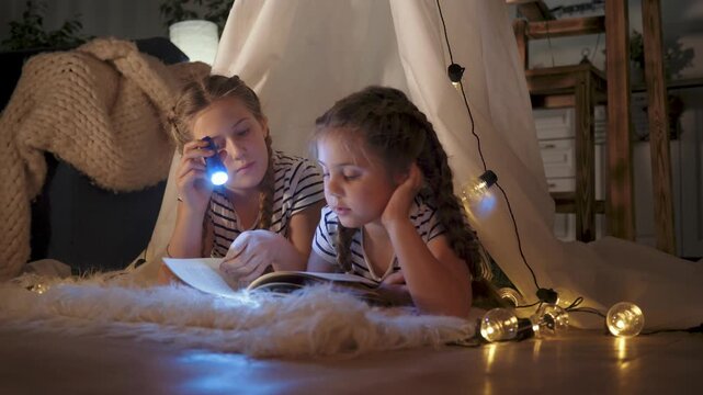 Girls play under blanket tent with flashlight sisters lie on rug using tablet pillow blanket and fairy light string create cozy indoor bedtime atmosphere with soft glow imaginative reading and sharing