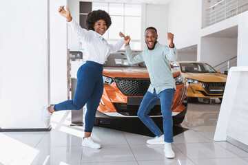 Young black couple cheering and dancing, gesturing YES, celebrating purchase of new car at auto dealership. Full length of Afro family buying automobile, feeling excited at showroom store