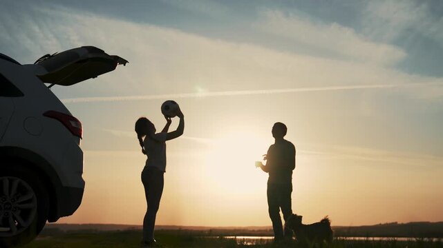 Sunset silhouettes play catch beside car trunk by lake and field while couple share relaxed travel break with ball between them warm sky reflection on water cloud streaks creating calm outdoor view