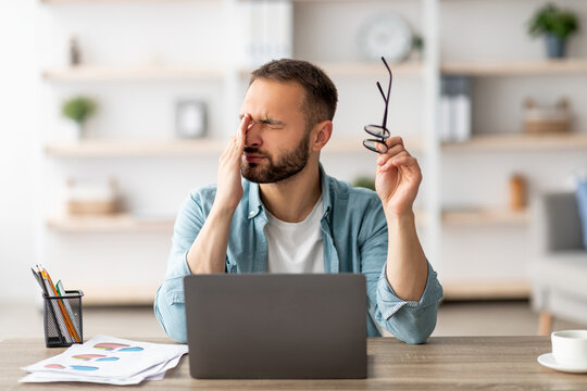 Caucasian male freelancer feeling tired, rubbing irritated eyes, sitting at desk with laptop, exhausted from online work. Young man having visual fatigue, doing business project on web