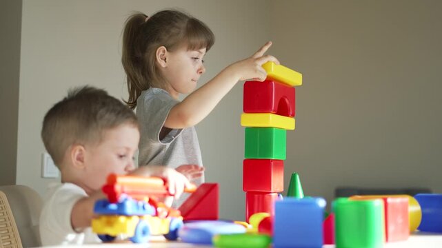 Child stacking colorful block tower with girl and boy playing at table, toy car and cup nearby, stacking block pieces in play and learning interaction, preschool activity showing focus and play.