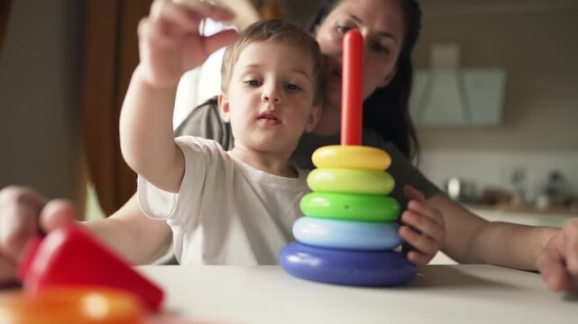 Mother helps toddler stack toy ring while parent guides child in learning play at kitchen table with colorful stacking ring set fostering fine motor skill and family bond through gentle interaction