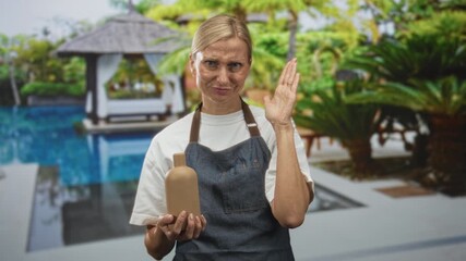 Woman holding a clay bottle with both hands while raising her right hand to ear and wearing an apron in a studio demonstration space; playful craft.