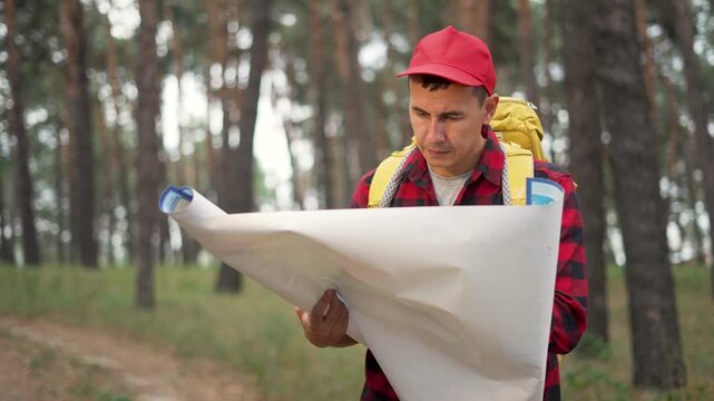 Reading map in dense pine forest man hiker wearing red cap and plaid shirt studies large map beside trail with yellow backpack on shoulder navigation for remote outdoors route planning and waypoint