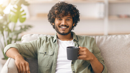 A man with curly hair sits on a couch in a living room. He smiles while holding a black cup....