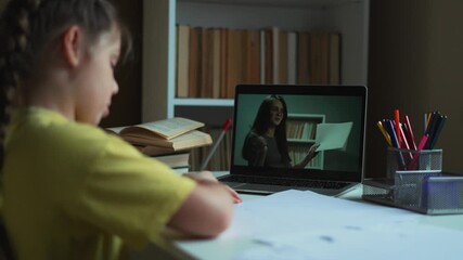 Child studies at desk with laptop and tutor on screen while girl writes homework using pencil and school book for study and online learning in quiet room with pen cup open notebook and lamp.